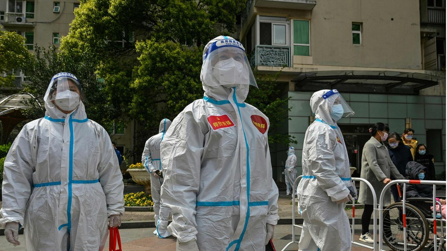 Drei Menschen in weißen Schutzanzügen sowie mit Gesichtsschild und FFP2-Masken schauen im Freien in unterschiedliche Richtungen
