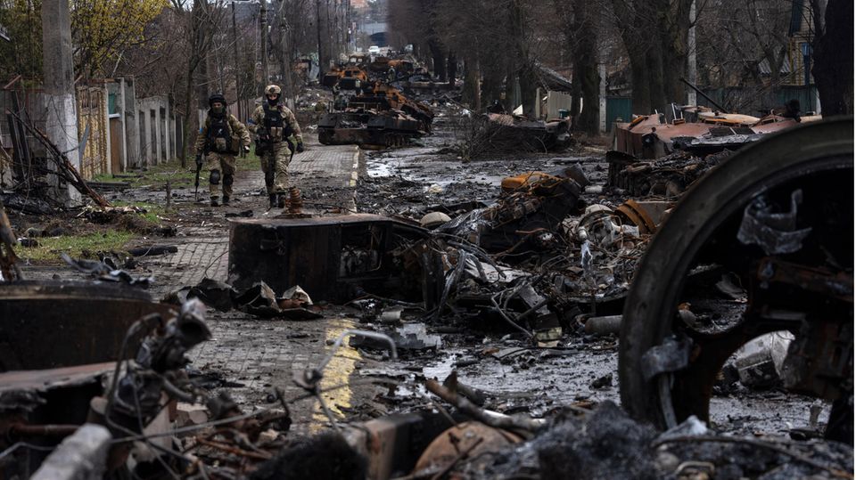 Zwei ukrainische Soldaten gehen auf einer Straße, die übersät ist mit zerstörten russischen Militärfahrzeugen