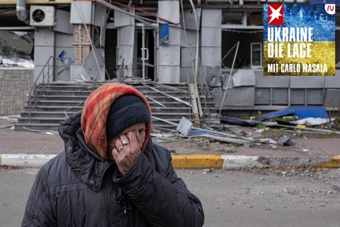 Eine alte Frau mit Kopftuch, Mütze und Winterjacke wischt sich vor einem beschädigten Haus mit links Tränen aus den Augen