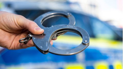 Eine Hand hält Handschellen vor einen Streifenwagen der Polizei