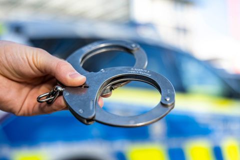 Eine Hand hält Handschellen vor einen Streifenwagen der Polizei