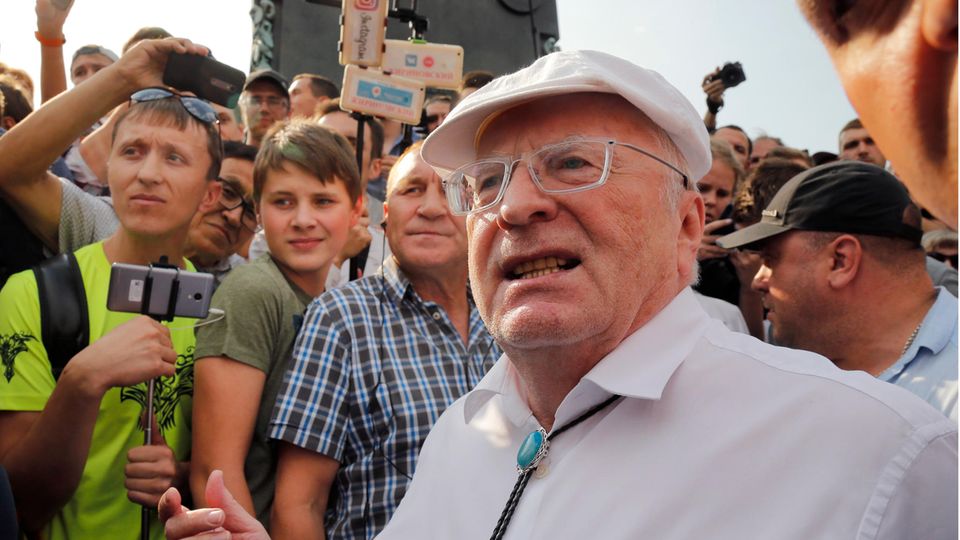 Ein alter weißer Mann mit Brille und weißer Schiebermütze steht in einer Menge junger Menschen