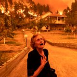 Eine alte Frau in schwarz greift sich an die Brust, während sich im Hintergrund ein Waldbrand Häusern nähert