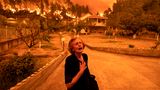 Eine alte Frau in schwarz greift sich an die Brust, während sich im Hintergrund ein Waldbrand Häusern nähert