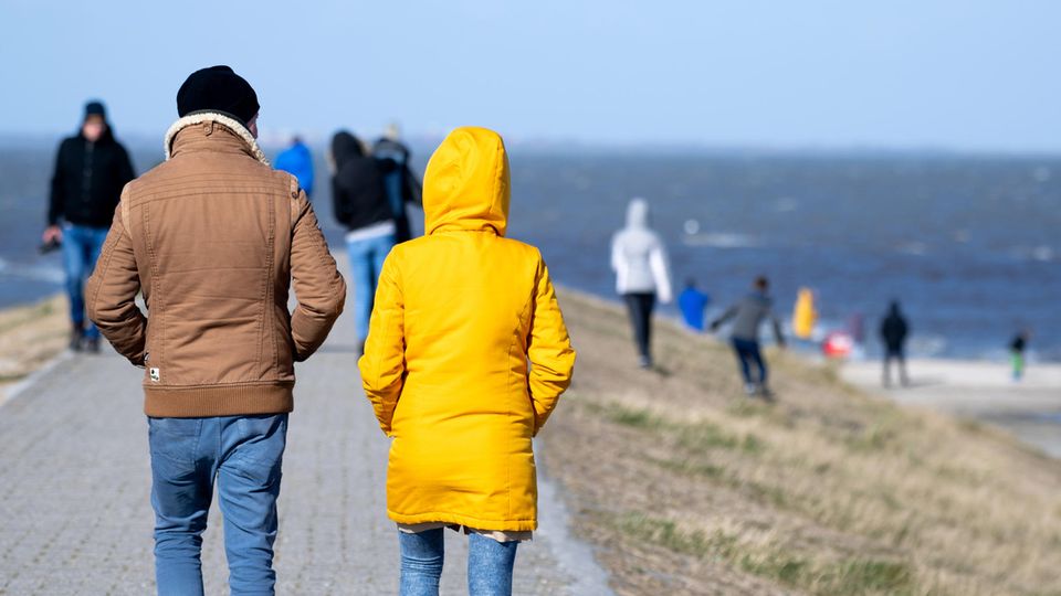 Touristen gehen bei sonnigem Wetter auf einem Deich am Hafen entlang.