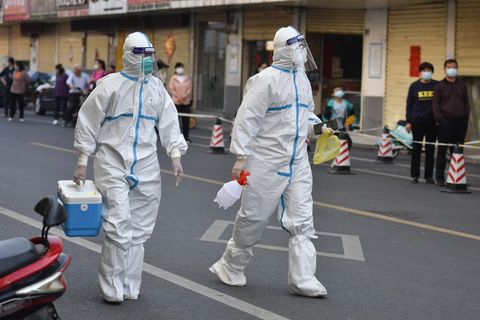 Menschen in weißen Overalls mit FFP2-Masken und Gesichtsschilden gehen auf einer Straße durch eine Großstadt