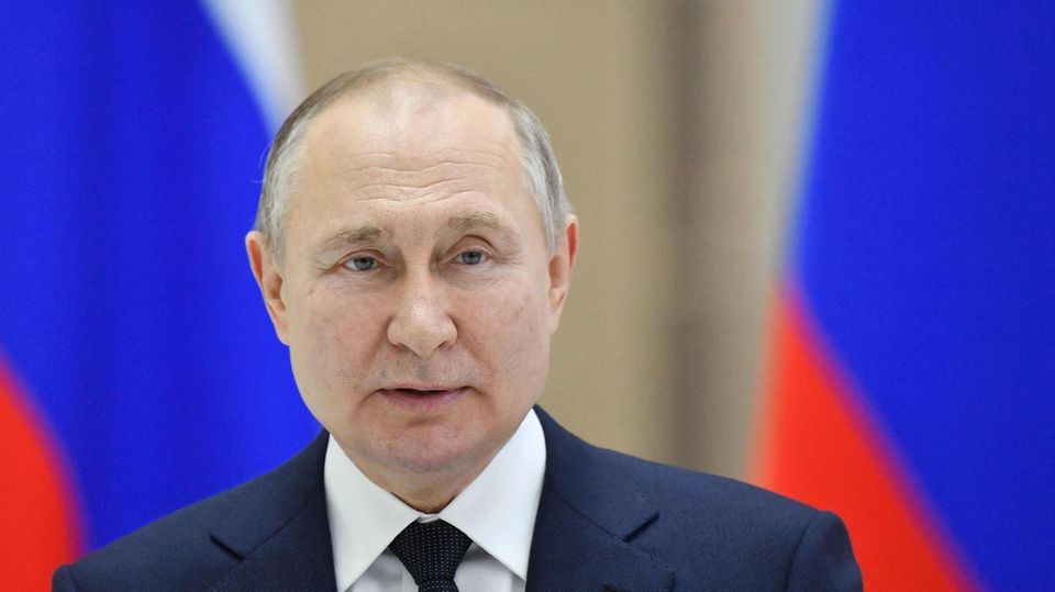 Wladimir Putin Ein weißer Mann mit rundlichem Gesicht und schütterem Haar steht in Anzug und Krawatte vor russischen Flaggen