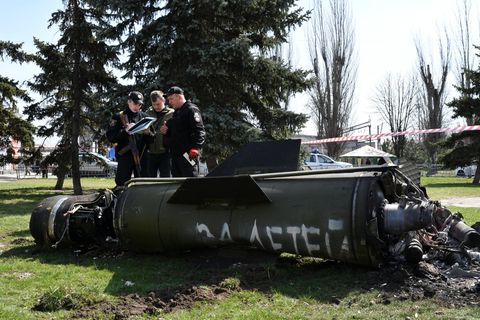 Hinter einem grünen Raketenteil stehen drei Männer in Uniformen und mit Klemmbrettern