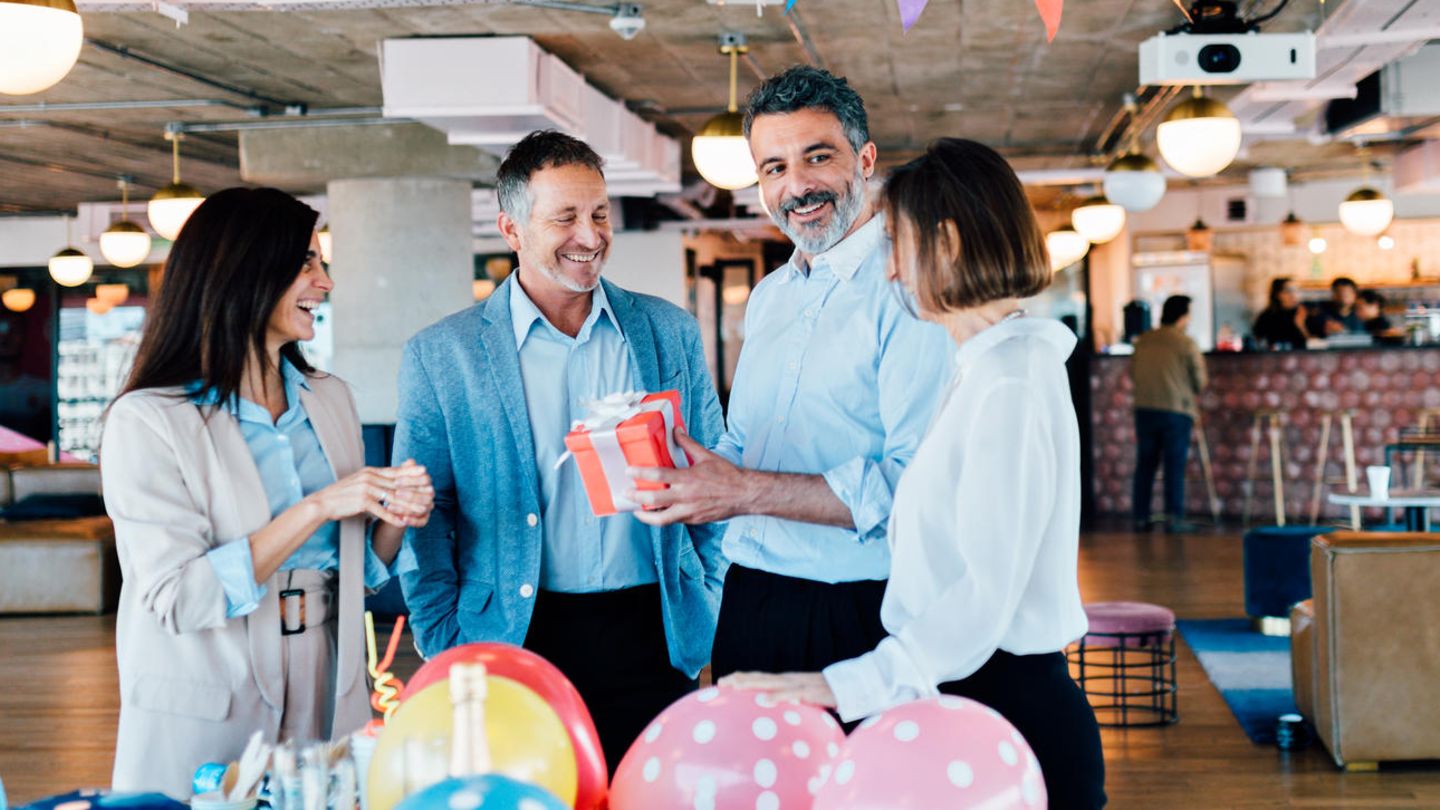 Vier Erwachsene stehen unter einer bunten Girlanden und hinter einem Tisch voller bunter Luftballons in einem Büro