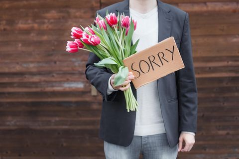Ein Mann hat einen Strauß Tulpen udn ein Schild mit dem Wort "Sorry" in der Hand