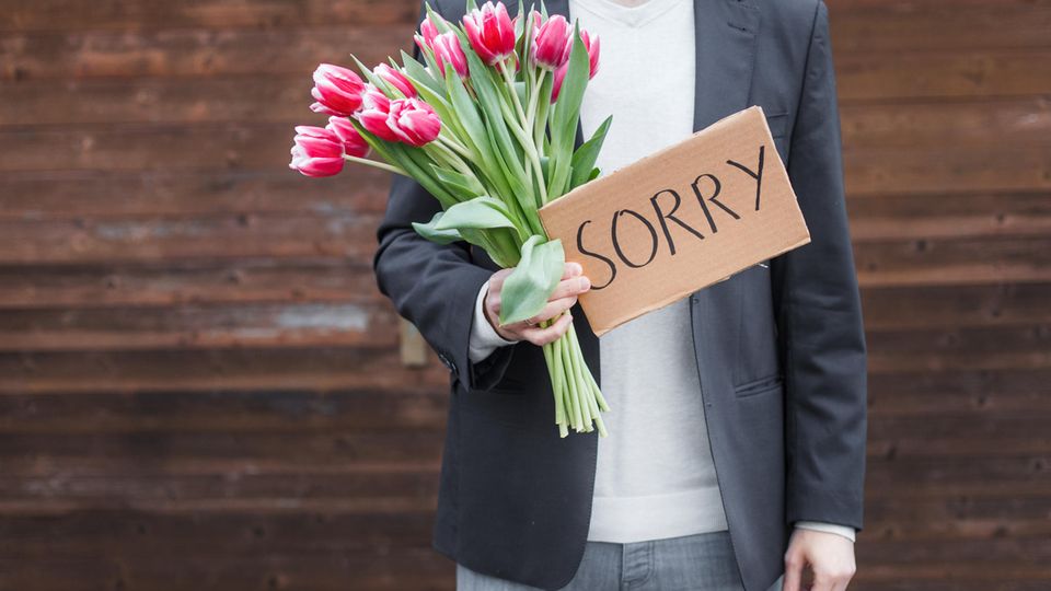Ein Mann hat einen Strauß Tulpen udn ein Schild mit dem Wort "Sorry" in der Hand