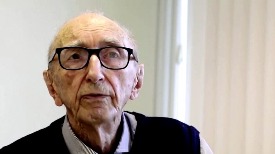Walter Orthmann Ein dünner, alter Mann mit schwarzem Brillengestell steht in einem hellen Büroraum