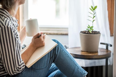 Frau sitzt mit einem Notizbuch und eine Tasse am Fenster