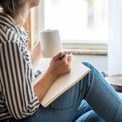 Frau sitzt mit einem Notizbuch und eine Tasse am Fenster