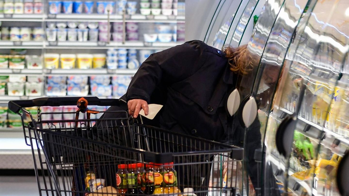 Eine Frau im Supermarkt beugt sich neben ihrem Einkaufswagen in ein Kühlregal, um einen Artikel zu greifen