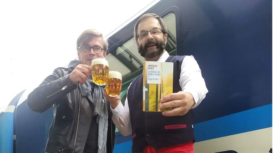Zugfahren verbindet: Autor Jaroslav Rudiš und Ober Pavel Peterka aus dem Speisewagen stoßen am Gleis mit Bier an