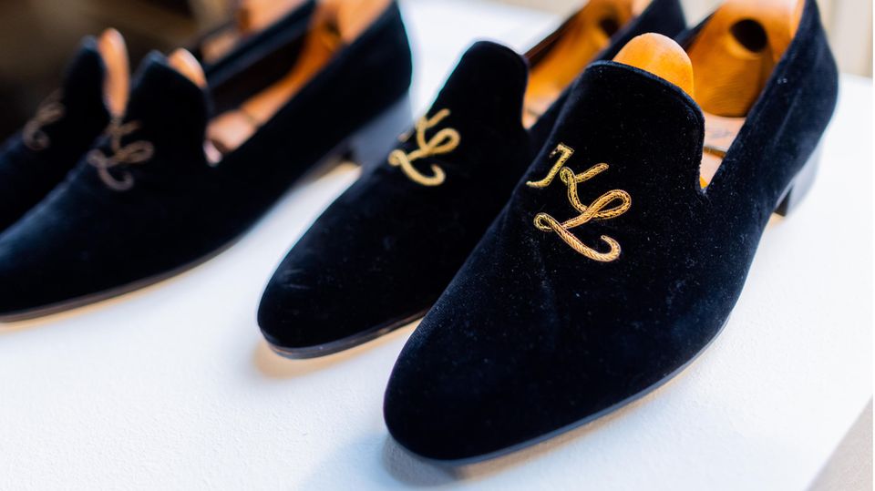 Blaue Samt-Slipper mit goldenem "KL"-Monogramm stehen auf einem weißen Podest