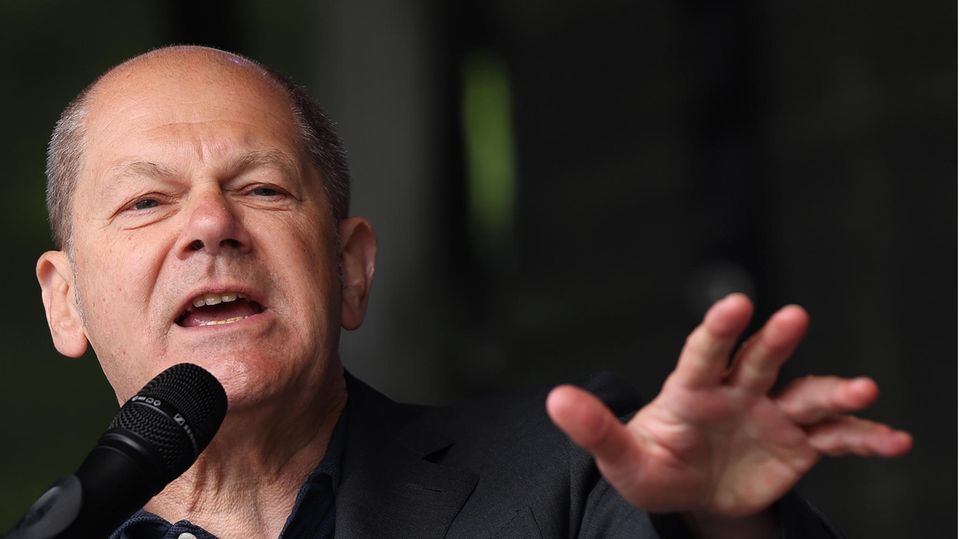 Ein älterer weißer Mann mit Halbglatze streckt seine linke Hand Richtung Publikum, während er auf einer Bühne eine Rede hält