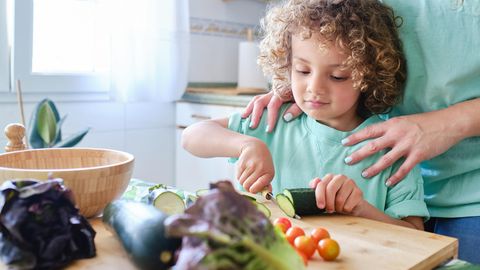 Kind steht it seiner Mutter in der Küche und schneidet Gemüse