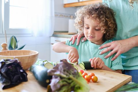 Kind steht it seiner Mutter in der Küche und schneidet Gemüse
