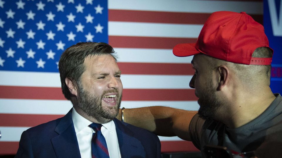 J.D. Vance Ein junger weißer Mann in Anzug und Krawatte lacht mit offenem Mund vor einer riesigen US-Flagge