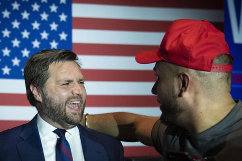 Ein junger weißer Mann in Anzug und Krawatte lacht mit offenem Mund vor einer riesigen US-Flagge