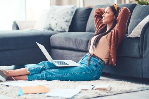 Eine junge Frau sitzt mit Laptop vor der Couch und schaut entspannt in den Raum