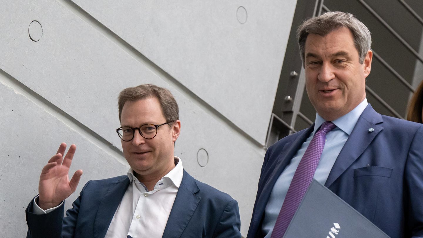 Zwei weiße Männer in Anzügen gehen nebeneinander eine Treppe herunter. Der linke winkt mit der rechten Hand