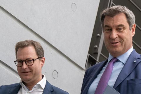 Zwei weiße Männer in Anzügen gehen nebeneinander eine Treppe herunter. Der linke winkt mit der rechten Hand