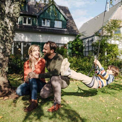 Ein Junge schaukelt im Garten, neben ihm seine Eltern