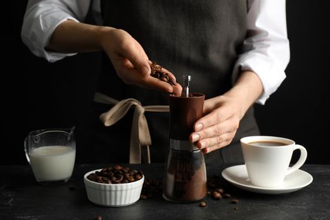 Handkaffeemühlen: Frau gibt an Tisch mit Tasse und Milche Kaffeebohnen in eine Kaffeemühle