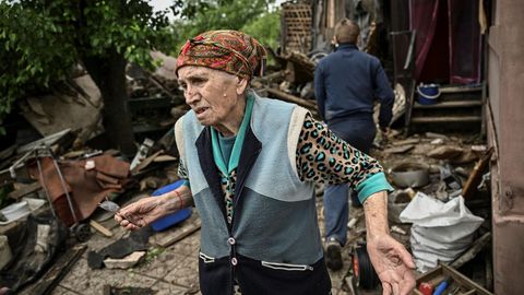 Eine alte Frau mit Kopftuch steht mit klagender Miene vor den Trümmern ihres einstigen Häuschens