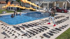 Ein Waffensammler steht vor einem Pool, umgeben von seinen Gewehren und hält einen Flammenwerfer hoch