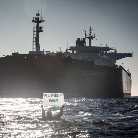 Im Gegenlicht hält ein Mensch im Wasser ein Banner "Oil fuels war" hoch, während ein riesiger Öltanker auf ihn zufährt