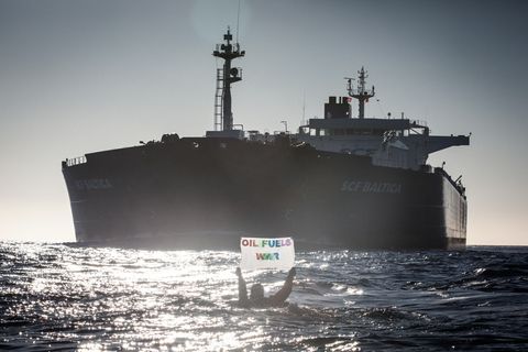 Im Gegenlicht hält ein Mensch im Wasser ein Banner "Oil fuels war" hoch, während ein riesiger Öltanker auf ihn zufährt
