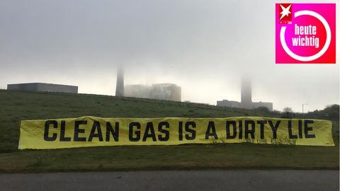 "Clean Gas is a dirty lie" steht in schwarz auf einem gelben Banner vor rauchenden Schornsteinen