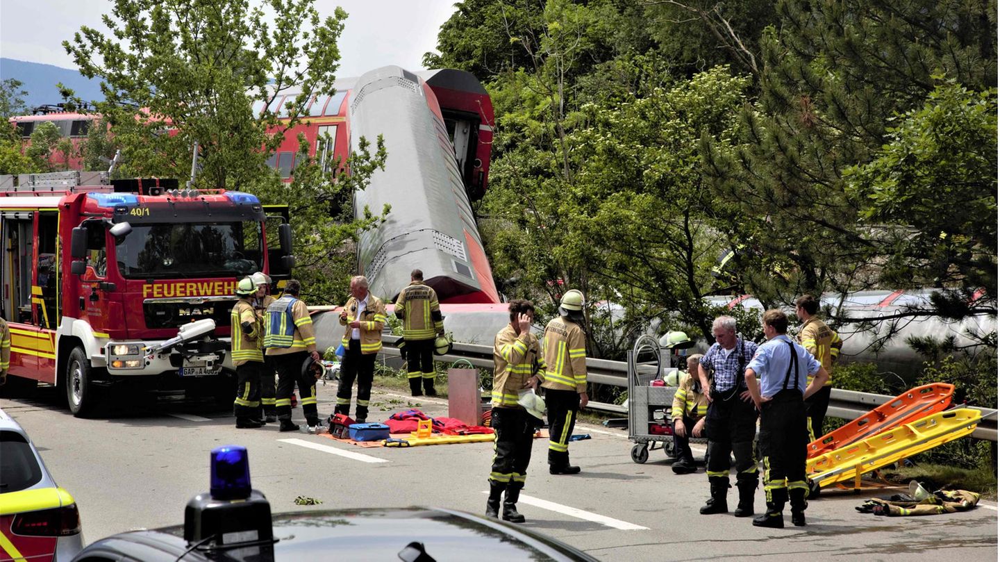 Feuerwehrmänner stehen an der Unglücksstelle, zahlreiche Opfer müssen aus den entgleisten Waggons geborgen werden. Zwei Waggons am Ende des Zugs liegen wie umgeworfen neben den Gleisen an einem kleinen Abhang.