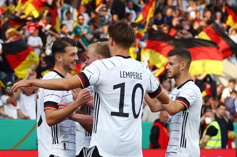 Vier junge Fußballspieler in weißen Trikots und schwarzen Hosen gehen mit ausgebreiteten Armen aufeinander zu und jubeln