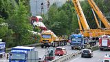 Im Morgenlicht stehen zwei Schwerlastkräne auf einer mehrspurigen Bundesstraße neben dem Gleis mit dem verunglückten Zug