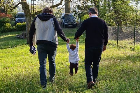 Patrick und Til halten ihre kleine Tochter auf einer Wiese an den Händen. Das schwule Paar hat das Mädchen adoptiert.