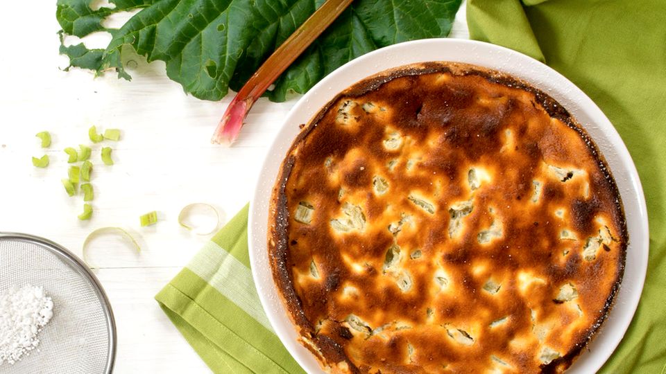 Die Mischung macht’s – Cremig-süßer Käsekuchen mit säuerlichem Rhabarber Kuchen und Rhabarber-Blätter, Puderzuckersieb mit Puderzucker auf weißem Holz