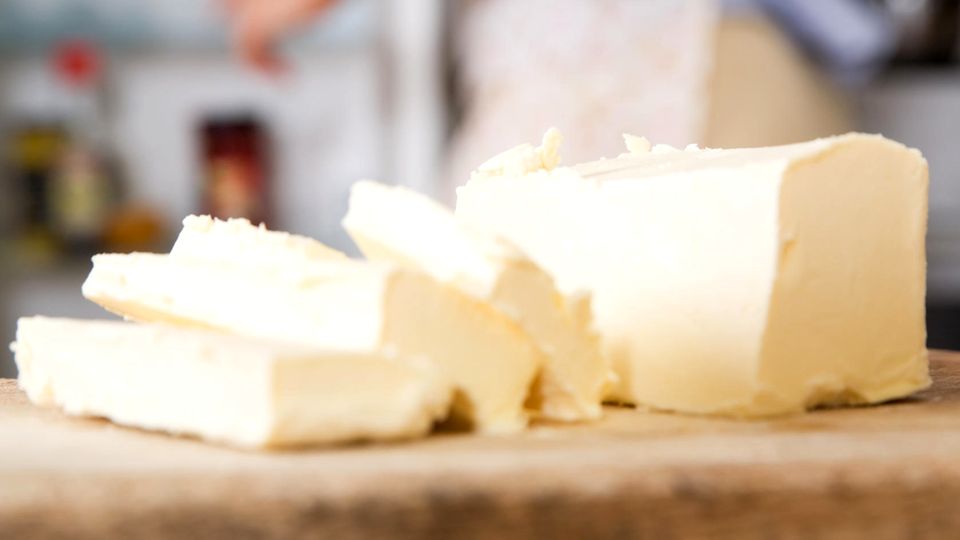 Butterpreise steigen – So können Sie sich Butter ganz einfach selbst machen Nahaufnahme von Butterscheiben, im Hintergrund Hausfrau öffnet den Kühlschrank.