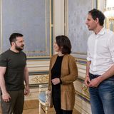 Ein weißer Mann in olivgrünem T-Shirt und Hose steht einer weißen Frau mit braunem Blazer und einem weißen Mann gegenüber.