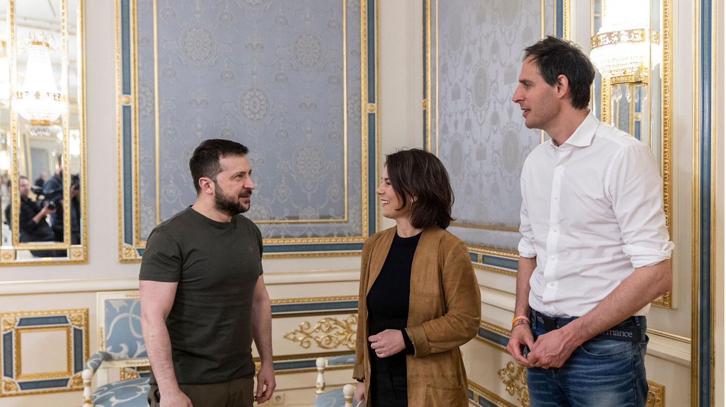 Ein weißer Mann in olivgrünem T-Shirt und Hose steht einer weißen Frau mit braunem Blazer und einem weißen Mann gegenüber. Ein weißer Mann in olivgrünem T-Shirt und Hose steht einer weißen Frau mit braunem Blazer und einem weißen Mann gegenüber.