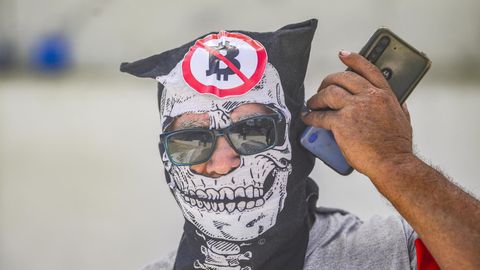 Ein Demonstrant telefoniert, während er während des Protestmarsches eine Maske mit Anti-Bitcoin-Symbolen trägt