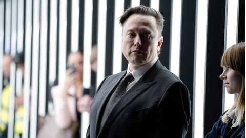 Elon Musk schaut mit ernstem Blick zur Seite, im Hintergrund sind Tesla-Mitarbeiter zu sehen