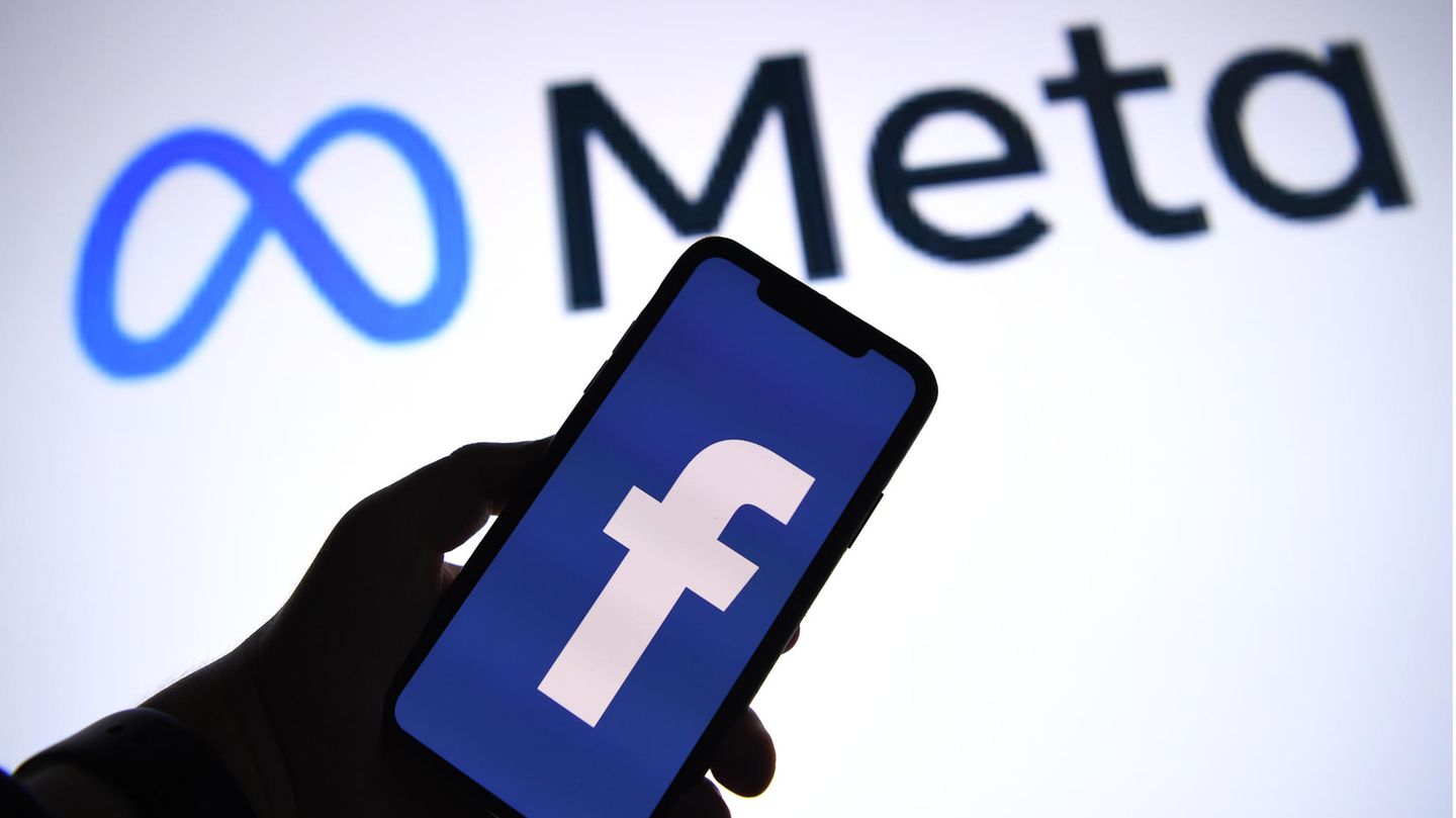 Eine Hand hält ein Smartphone mit der geöffneten Facebook-App hoch, im Hintergrund der Schriftzug "Meta"