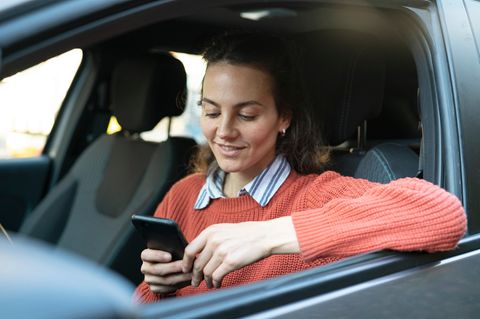 Eine junge Frau sitzt am Steuer und sucht auf ihrem Smartphone nach Tankstellen