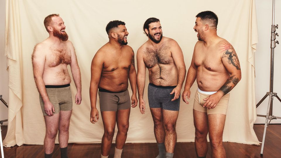 Vier hemdlose Männer lachen fröhlich, während sie vor einem Studiohintergrund stehen