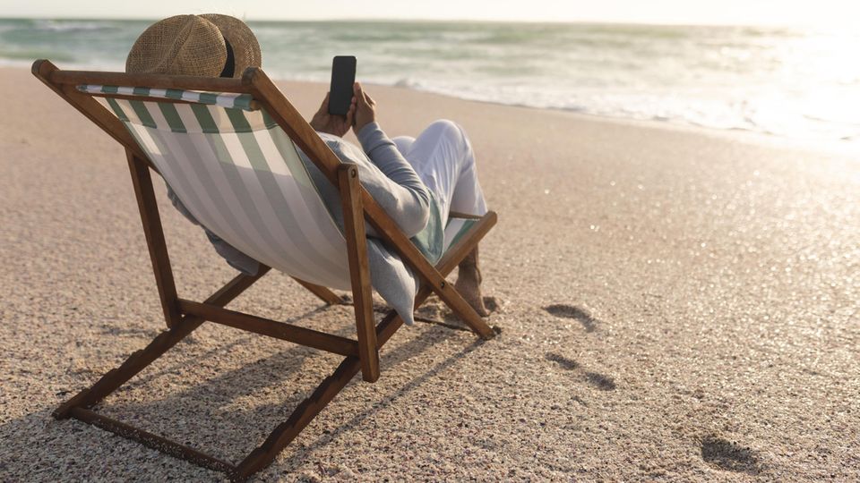 A man lies on a deck chair on the beach with a smartphone